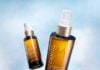 site-industries-cosmetiques Two brown Moroccanoil treatment bottles with white caps are shown against a sky-blue background. One bottle is upright and closer, while the other is angled and further away.