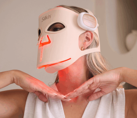 site-industries-cosmetiques Une personne portant un haut blanc sans manches et un masque de luminothérapie à LED dont la marque "Silk'n" est visible. Le masque émet une lumière rouge et la personne tient ses mains sous son menton.