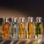 site-industries-cosmetiques Five glass perfume bottles by Bormioli Luigi with gold labels and crystal stoppers are arranged in rows on a brown surface with warm lighting, each featuring Spoturno-inspired glass innovations and containing a different-colored liquid.