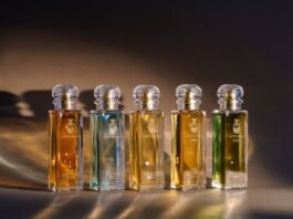 site-industries-cosmetiques Five glass perfume bottles by Bormioli Luigi with gold labels and crystal stoppers are arranged in rows on a brown surface with warm lighting, each featuring Spoturno-inspired glass innovations and containing a different-colored liquid.
