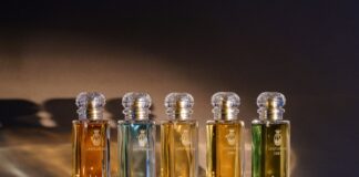 site-industries-cosmetics Cinco frascos de perfume de vidrio de Bormioli Luigi con etiquetas doradas y tapones de cristal se disponen en fila sobre una superficie marrón con una cálida iluminación, cada uno con innovaciones de vidrio inspiradas en Spoturno y conteniendo un líquido de distinto color.