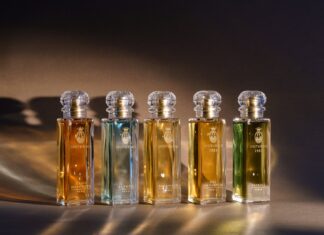 site-industries-cosmetiques Five glass perfume bottles by Bormioli Luigi with gold labels and crystal stoppers are arranged in rows on a brown surface with warm lighting, each featuring Spoturno-inspired glass innovations and containing a different-colored liquid.