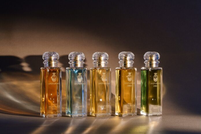 site-industries-cosmetics Cinco frascos de perfume em vidro da autoria de Bormioli Luigi, com rótulos dourados e rolhas de cristal, estão dispostos em fila numa superfície castanha com iluminação quente, cada um deles com inovações em vidro inspiradas em Spoturnos e contendo um líquido de cor diferente.