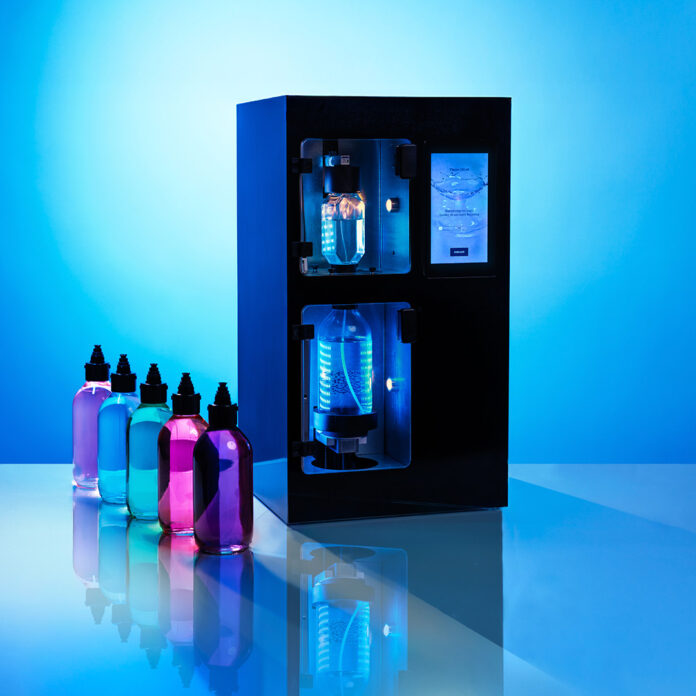 site-industries-cosmetiques Uma máquina retangular preta com duas câmaras visíveis, iluminadas por uma luz azul. Quatro garrafas de plástico cheias de líquidos de cores diferentes - rosa, azul, verde e violeta - estão dispostas à esquerda sobre uma superfície reflectora.