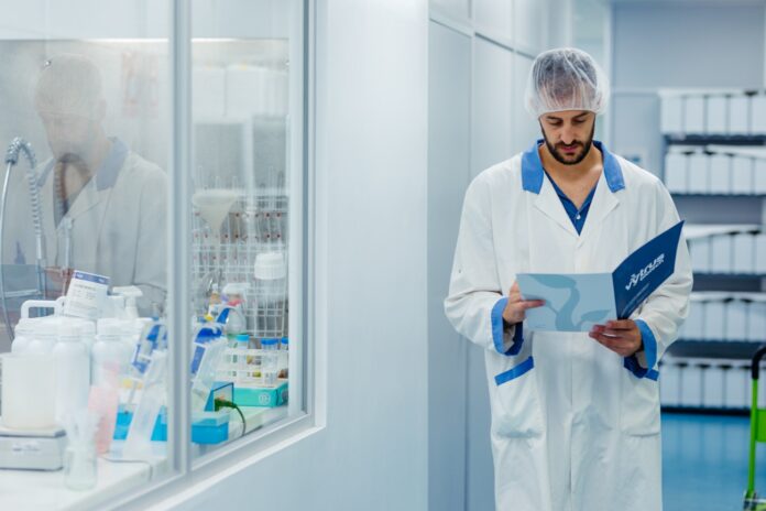 site-industries-cosmetiques Um homem vestindo uma bata branca e uma rede de pesca está a ler um documento num corredor de um laboratório. O equipamento de laboratório e os frascos são visíveis através de uma janela à sua esquerda.