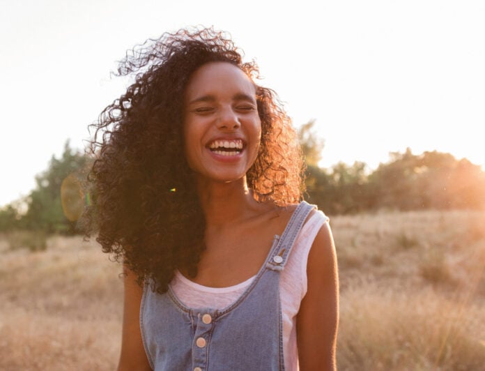 site-industries-cosmetiques Una mujer con el pelo rizado sonríe y ríe al aire libre en un campo soleado, vestida con un top blanco y un mono vaquero. Al fondo se ven árboles y hierba alta.