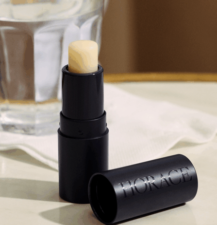 site-industries-cosmetiques Um tubo preto de bálsamo labial com o nome da marca "HORACE" gravado na tampa, colocado numa superfície de mármore ao lado de um copo de água e de um guardanapo branco.
