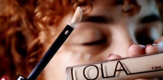 site-industries-cosmetiques 一位卷发女士正拿着一管标有 "LOLA "字样的遮瑕膏贴近自己的脸，并在眼睛下方涂抹了一点遮瑕膏。她闭着眼睛，似乎在微微笑。.