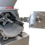 site-industries-cosmetiques A stainless steel industrial milling machine with open front panel, revealing internal blades and a perforated curved sieve. The detached lid is hinged to the side.