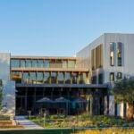 site-industries-cosmetiques Um edifício moderno com grandes janelas, revestimento metálico e linhas simples, rodeado por árvores jovens e vegetação paisagística sob um céu azul claro. Várias mesas ao ar livre com guarda-sóis podem ser vistas perto da entrada.