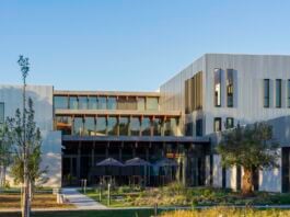 site-industries-cosmetiques Un bâtiment moderne avec de grandes fenêtres, un bardage métallique et des lignes épurées, entouré de jeunes arbres et de verdure paysagère sous un ciel bleu clair. Plusieurs tables extérieures avec parasols sont visibles près de l'entrée.