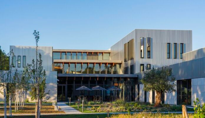 site-industries-cosmetiques Un bâtiment moderne avec de grandes fenêtres, un bardage métallique et des lignes épurées, entouré de jeunes arbres et de verdure paysagère sous un ciel bleu clair. Plusieurs tables extérieures avec parasols sont visibles près de l'entrée.