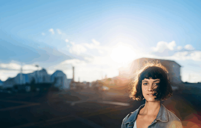 site-industries-cosmetiques Uma mulher com cabelo castanho curto e encaracolado está lá fora, à luz do sol. Veste um casaco de ganga e o fundo mostra uma rua vazia, edifícios e um céu claro e brilhante com algumas nuvens.