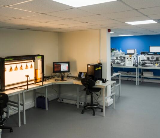site-industries-cosmetiques Un laboratoire propre et moderne comprenant des postes de travail, un équipement Dia-Stron pour l'analyse visuelle des cheveux, des ordinateurs et des imprimantes 3D. L'espace est composé de murs blancs, d'un mur d'accent bleu, d'un éclairage fluorescent au plafond et de deux chaises de bureau.