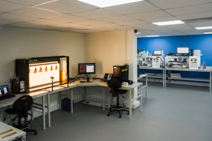 Lab image 3 site-industries-cosmetiques Un laboratoire propre et moderne comprenant des postes de travail, un équipement Dia-Stron pour l'analyse visuelle des cheveux, des ordinateurs et des imprimantes 3D. L'espace est composé de murs blancs, d'un mur d'accent bleu, d'un éclairage fluorescent au plafond et de deux chaises de bureau.