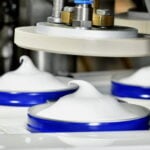 site-industries-cosmetiques Grande plano de uma máquina industrial automática Brouillon que distribui natas batidas ou uma substância branca semelhante em recipientes redondos azuis, formando um remoinho pontiagudo no topo de cada um deles.