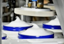 site-industries-cosmetiques 一台布劳永汽车工业机器的特写，该机器将鲜奶油或类似的白色物质分装到圆形的蓝色容器中，在每个容器的顶部形成一个尖形漩涡。
