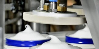 site-industries-cosmetiques Gros plan d'une machine industrielle Brouillon auto distribuant de la crème fouettée ou une substance blanche similaire dans des récipients ronds bleus, formant un tourbillon pointu sur le dessus de chacun d'entre eux.