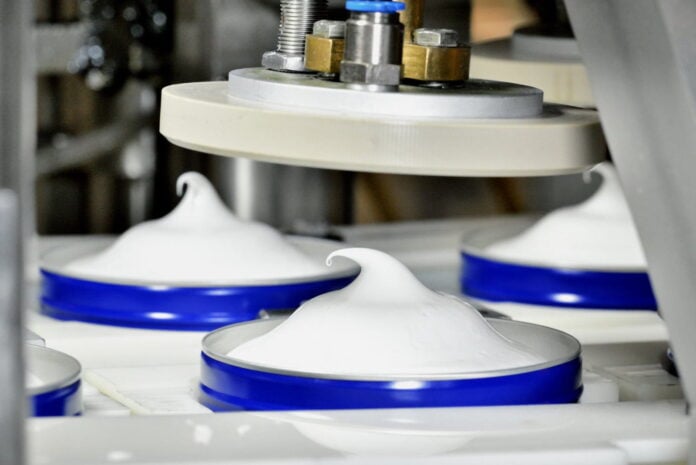 site-industries-cosmetiques Grande plano de uma máquina industrial automática Brouillon que distribui natas batidas ou uma substância branca semelhante em recipientes redondos azuis, formando um remoinho pontiagudo no topo de cada um deles.