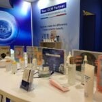 ALPOL Cosmétique 的创新季 site-industries-cosmetiques 展台上展示着各种瓶装、罐装和管装护肤品和化妆品,并配有信息板。背景中的蓝白条幅展示了 ALPOL Cosmétique 作为 "您的 OEM 合作伙伴 "的形象,突出了美容产品生产中的创新。.
