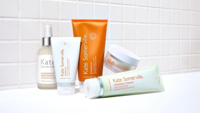 site-industries-cosmetiques Cinq produits de soin de la peau de Kate Somerville, de la crème à l'exfoliant, sont présentés sur un fond de carreaux blancs. Cette collection Rare Beauty Brands présente des emballages blancs et orange, clairement étiquetés avec le nom de confiance d'Unilever.