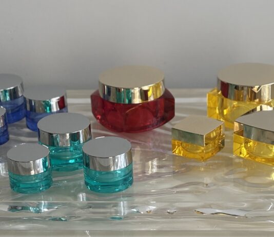 site-industries-cosmetics 蓝色、红色和黄色的各种塑料化妆品罐和容器，配以银色盖子，摆放在质感透明的表面上，背景为纯灰色。.