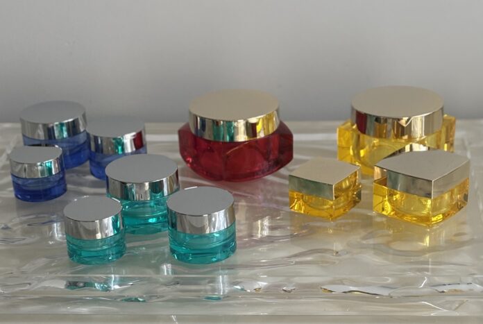 site-industries-cosmetics 蓝色、红色和黄色的各种塑料化妆品罐和容器，配以银色盖子，摆放在质感透明的表面上，背景为纯灰色。.