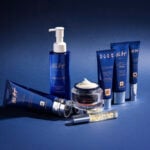 site-industries-cosmetiques Une gamme courte de six produits de soins dermo-cosmétiques bleus Pier Augé - flacons de lotion, pots de crème et tubes - est disposée sur une surface bleue avec un fond assorti. Un pot est ouvert, révélant un tourbillon de crème blanche.