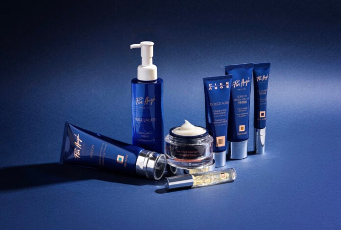site-industries-cosmetiques Une gamme courte de six produits de soins dermo-cosmétiques bleus Pier Augé - flacons de lotion, pots de crème et tubes - est disposée sur une surface bleue avec un fond assorti. Un pot est ouvert, révélant un tourbillon de crème blanche.
