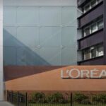 site-industries-cosmetiques L'extérieur d'un bâtiment moderne avec une grande enseigne "L'ORÉAL" fixée sur un mur de briques, à côté d'une façade en verre et d'un espace vert, reflétant la croissance et l'innovation de la marque.