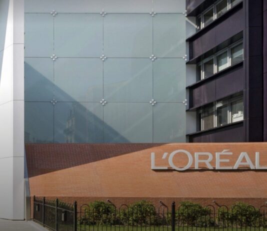 site-industries-cosmetiques L'extérieur d'un bâtiment moderne avec une grande enseigne "L'ORÉAL" fixée sur un mur de briques, à côté d'une façade en verre et d'un espace vert, reflétant la croissance et l'innovation de la marque.