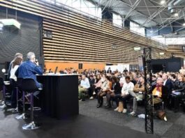 site-industries-cosmetiques Un panel de quatre personnes est assis à une table sur une scène face à un public dans une grande salle moderne. Le public est assis et écoute attentivement. L'espace a de hauts plafonds et des panneaux de bois rayés sur les murs.