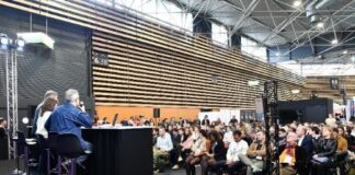 site-industries-cosmetiques Um painel de quatro pessoas está sentado a uma mesa num palco, de frente para o público, num grande salão moderno. O público está sentado e a ouvir atentamente. O espaço tem tectos altos e painéis de madeira às riscas nas paredes.