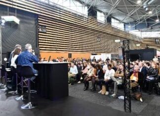 site-industries-cosmetiques Un panel de quatre personnes est assis à une table sur une scène face à un public dans une grande salle moderne. Le public est assis et écoute attentivement. L'espace a de hauts plafonds et des panneaux de bois rayés sur les murs.