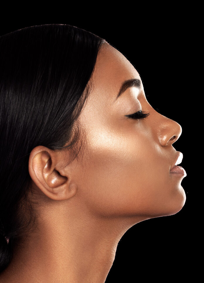 site-industries-cosmetiques Perfil lateral em grande plano de uma jovem mulher com cabelo preto e pele suave e radiante. Os seus olhos estão fechados e a cabeça ligeiramente levantada, realçando o maxilar e as maçãs do rosto num fundo preto.