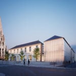 site-industries-cosmetiques Un bâtiment moderne en verre et un bâtiment traditionnel en pierre se trouvent côte à côte près d'une église historique en pierre dotée d'une haute flèche, en France. À la tombée de la nuit, plusieurs personnes marchent ou se tiennent debout sur la zone pavée, reflétant le mélange de patrimoine et d'innovation cosmétique de la ville.