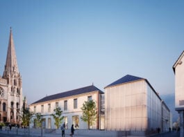 site-industries-cosmetiques Un bâtiment moderne en verre et un bâtiment traditionnel en pierre se trouvent côte à côte près d'une église historique en pierre dotée d'une haute flèche, en France. À la tombée de la nuit, plusieurs personnes marchent ou se tiennent debout sur la zone pavée, reflétant le mélange de patrimoine et d'innovation cosmétique de la ville.