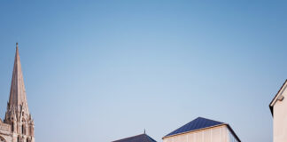 site-industries-cosmetiques 在法国，一座现代化的玻璃建筑和一座传统的石头建筑并排矗立在一座历史悠久、尖顶高耸的石头教堂旁。夜幕降临，许多人在鹅卵石铺就的街道上行走或站立，体现了这座小镇传统与化妆品创新的融合。