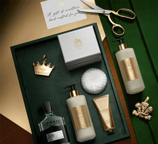 site-industries-cosmetiques 一套 Creed Aventus 豪华沐浴和身体护理产品，与 Kering 的优雅风格相呼应，包括一瓶古龙水、两个泵瓶、一个软管、一块香皂和一个金色标签，全部摆放在一个深色托盘上，背景是金色剪刀和一张纸条。