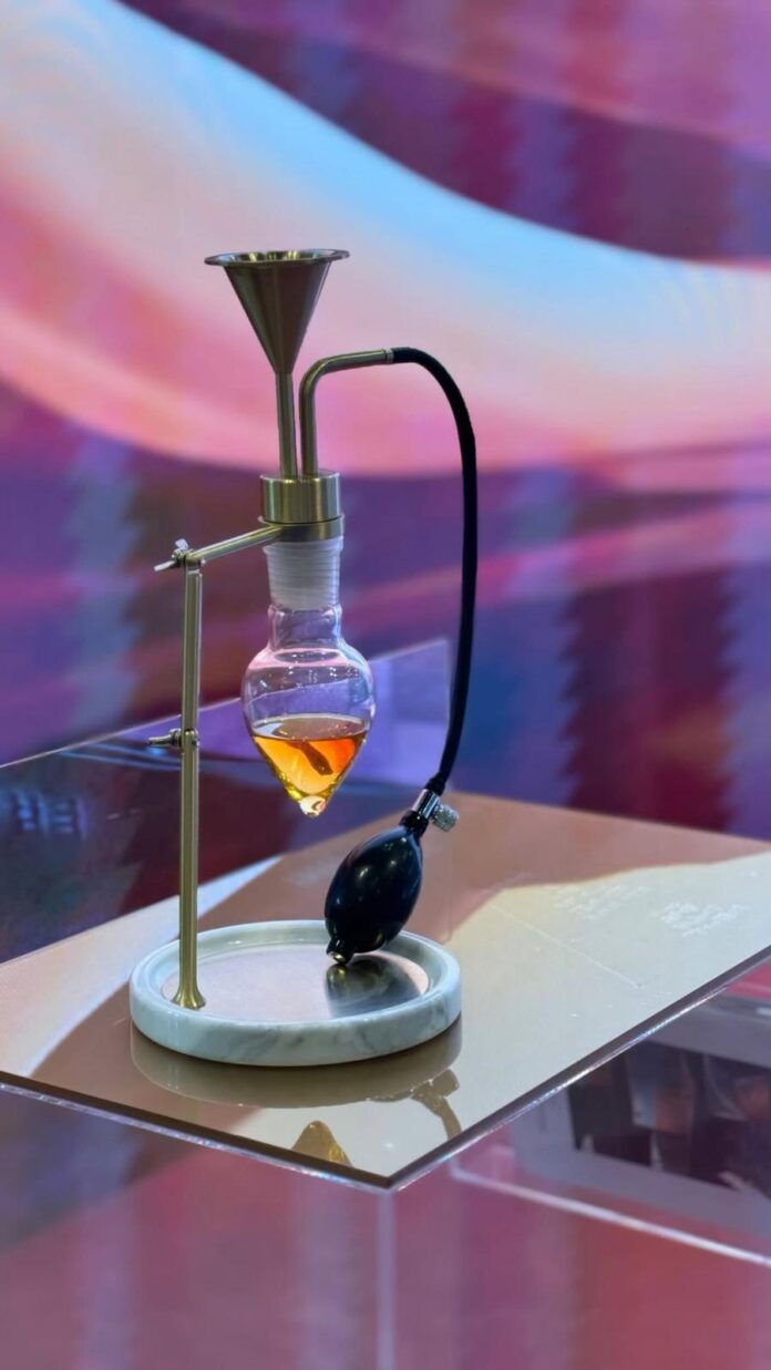 site-industries-cosmetiques EMSA Esencias Moles laboratory apparatus, comprising a glass ampoule containing an orange liquid, a metal support, a funnel and a black rubber ampoule connected by a tube - an innovative device for creating the characteristic Odor 2025 on a reflective surface.