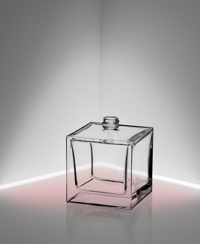 site-industries-cosmetiques Una botella de cristal cuadrada, transparente y vacía, con un pequeño cuello, se encuentra en la esquina de una habitación. El fondo y el suelo son blancos, con una tenue luz rosa que ilumina el suelo cerca de la base de la botella.