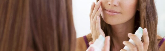 site-industries-cosmetiques Uma mulher está em frente a um espelho e aplica creme na bochecha com os dedos, segurando um frasco de cosméticos Silgan Dispensing numa mão. O seu reflexo é visível no espelho.