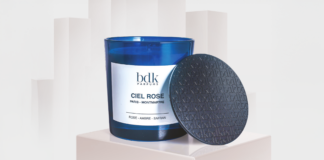site-industries-cosmetiques Une bougie en verre bleu étiquetée "bdk Parfums Ciel Rose" est posée sur un piédestal blanc à côté duquel se trouve un couvercle noir texturé. L'arrière-plan est légèrement flou dans des tons clairs.