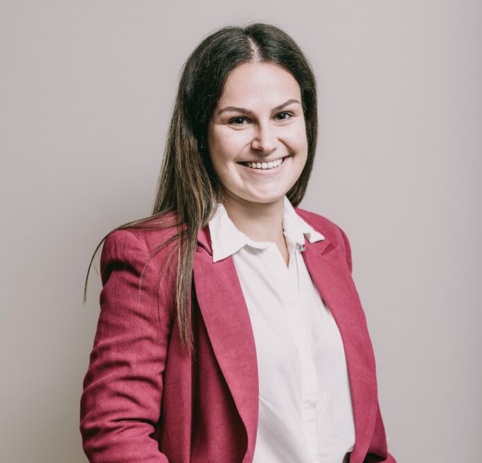 site-industries-cosmetiques Una mujer con el pelo largo y castaño lleva una camisa blanca y una americana roja. Sonríe y mira a la cámara, sobre un fondo liso de color claro.