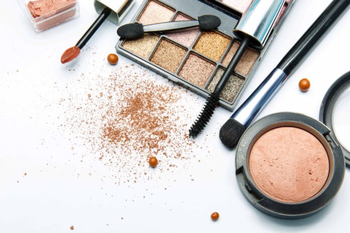 site-industries-cosmetiques Vários produtos de maquilhagem, incluindo uma paleta de sombras, um aplicador de batom, um blush compacto, um aplicador de rímel, pó facial e pó cosmético espalhados numa superfície branca.