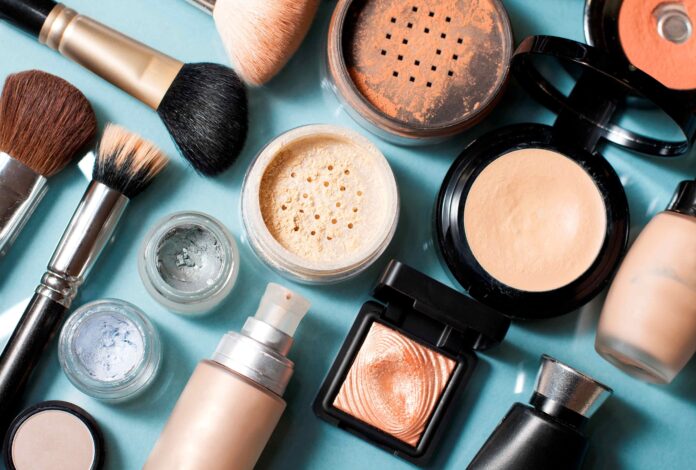 site-industries-cosmetiques Vários produtos de maquilhagem e pincéis estão expostos numa superfície azul clara. Os artigos incluem pós soltos e compactos, bases líquidas, marcadores, potes de sombras e vários pincéis de maquilhagem.