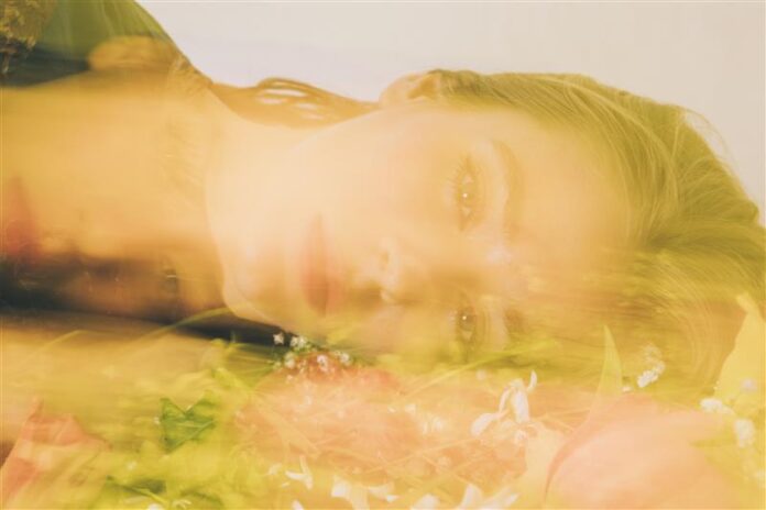 site-industries-cosmetiques A fair-skinned person with blond hair lies on the ground, surrounded by flowers. The image has a soft, yellowish blur effect, creating a dreamy, ethereal atmosphere.