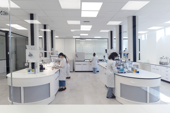 site-industries-cosmetiques Plusieurs personnes portant des blouses blanches travaillent séparément à différents postes dans un laboratoire moderne et lumineux équipé d'instruments scientifiques et de verrerie. De grandes fenêtres apportent de la lumière naturelle.