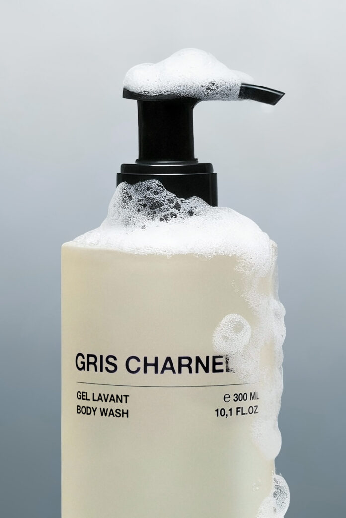 site-industries-cosmetiques Un frasco de gel de baño Gris Charnel con tapón de bomba, parcialmente cubierto de burbujas de jabón espumosas, sobre un fondo gris liso. La etiqueta indica un volumen de 300 ml.