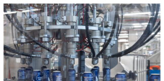 site-industries-cosmetiques Un primer plano de una cadena de producción industrial muestra el llenado de latas de aluminio azules por una máquina automatizada con múltiples boquillas y tubos negros, en un entorno industrial.